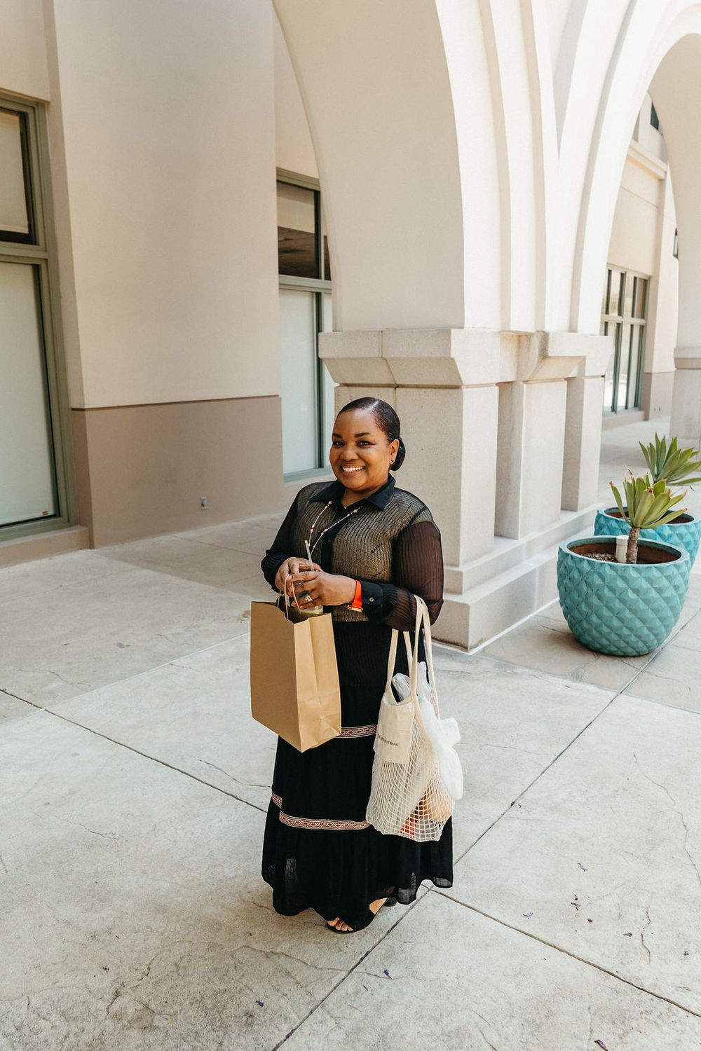 Smiling woman in black dress holding shopping bags outside a building with arched columns.