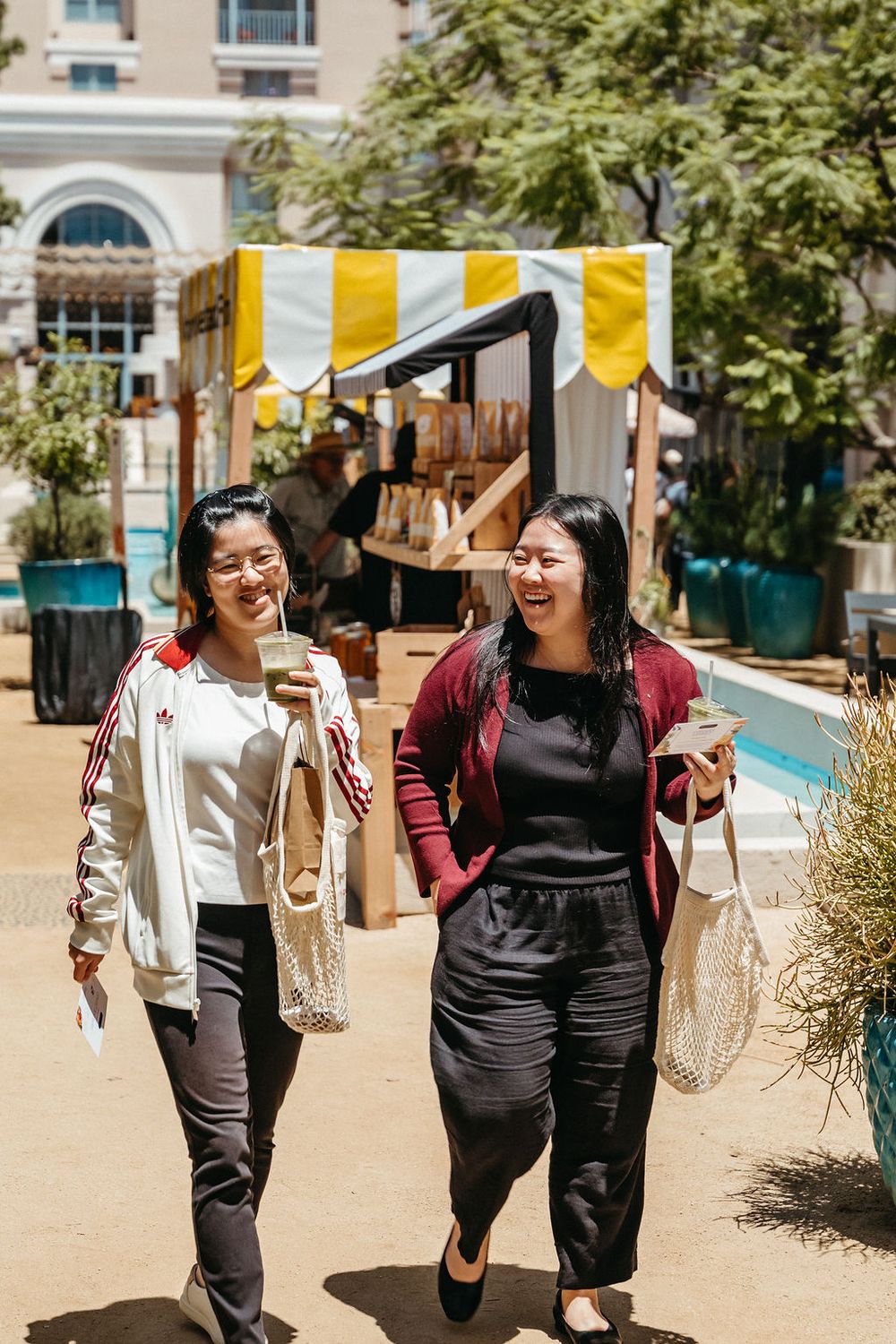 Two women laughing and walking at an outdoor market with yellow striped awning.