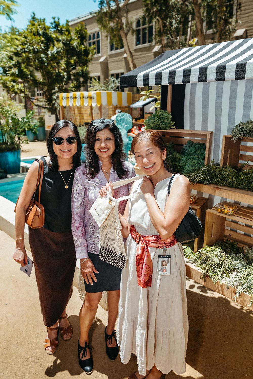 Three women smiling together at an outdoor market event with striped awnings and greenery in the background.