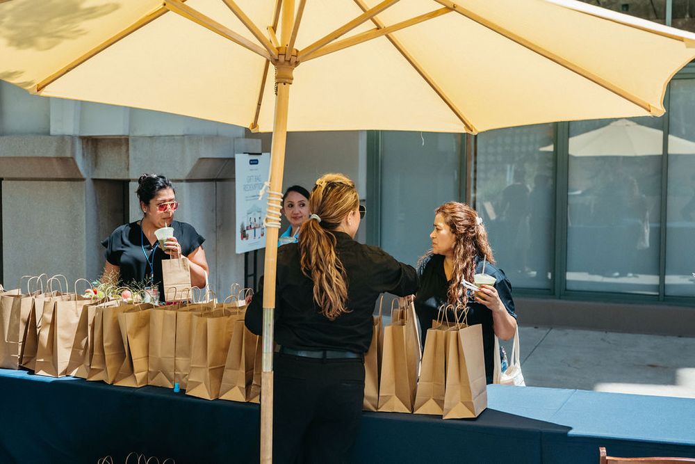 Women browsing kraft paper bags at an outdoor vendor table under a yellow umbrella.