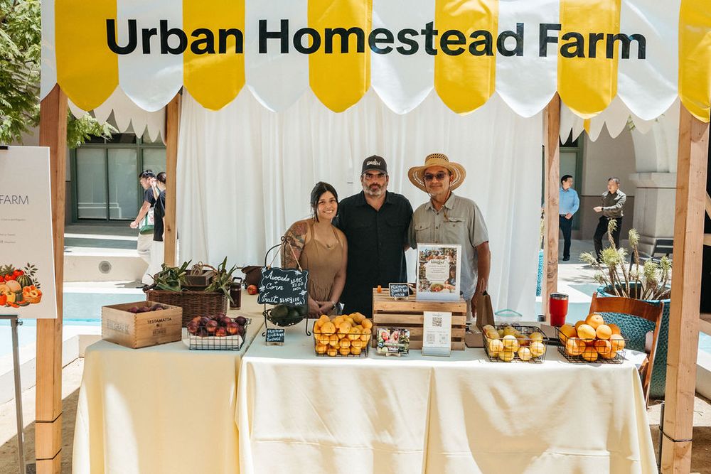 Three vendors smile at Urban Homestead Farm market booth with fresh produce and yellow balloon decorations.