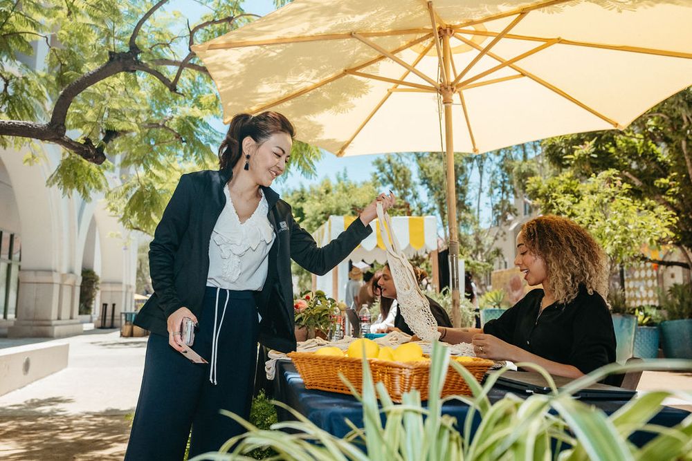 Woman receiving a drink from vendor at sunny outdoor market under yellow umbrella.