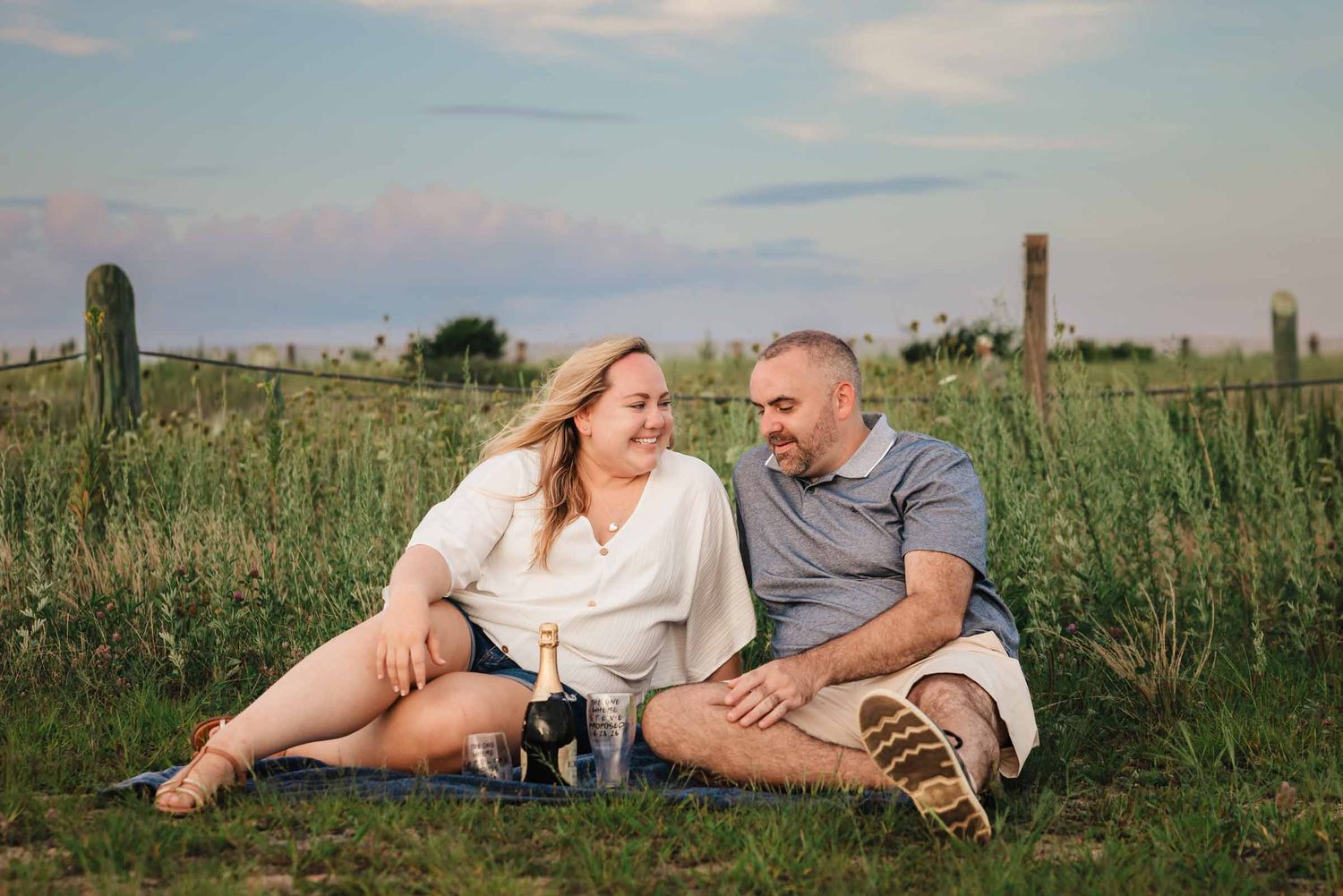 Engaged couple sharing a laugh during a picnic in a grassy field at Duxbury Beach, Massachusetts, with champagne and glasses.