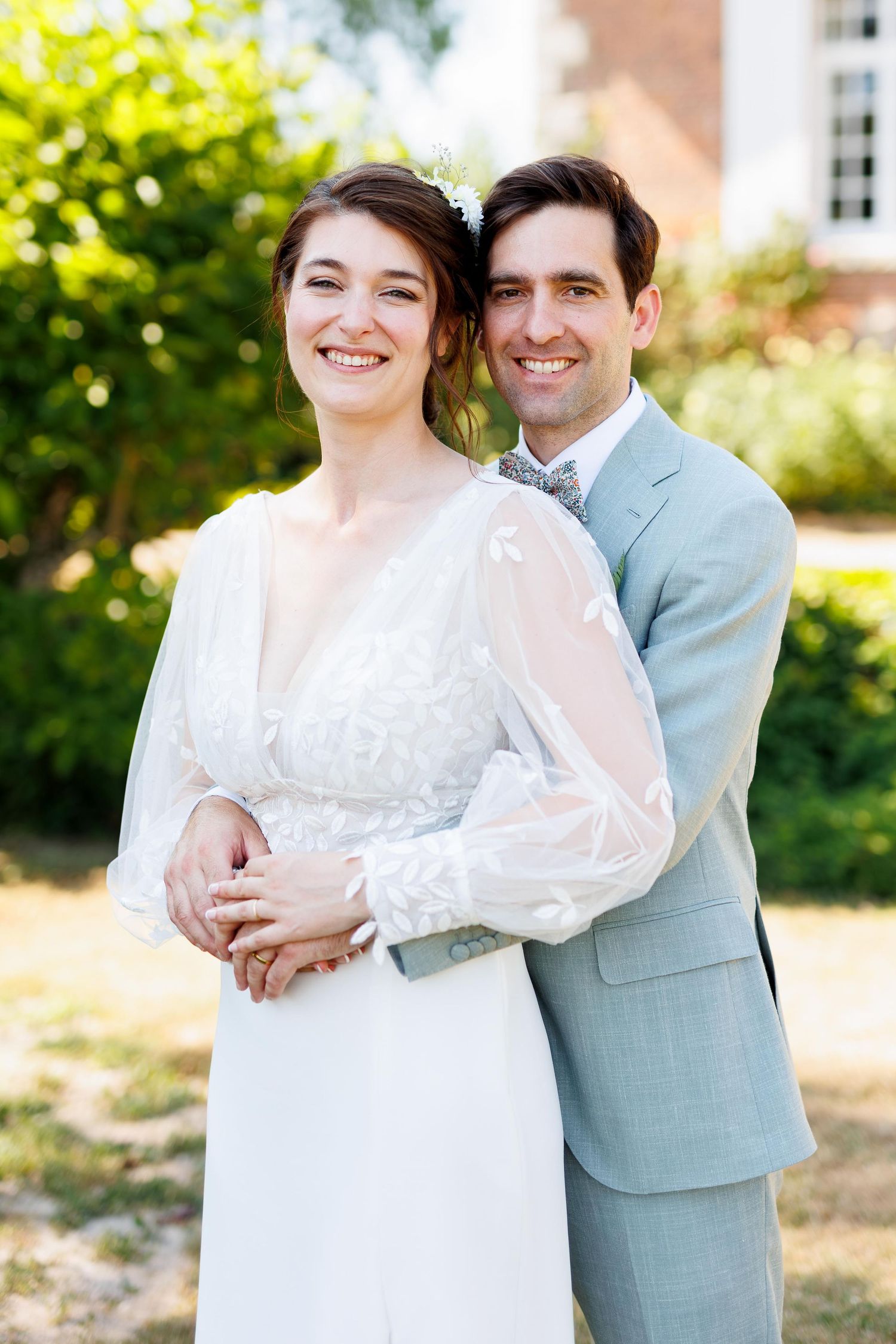 Bride in white gown holding greenery bouquet stands with groom in light gray suit outside brick estate.