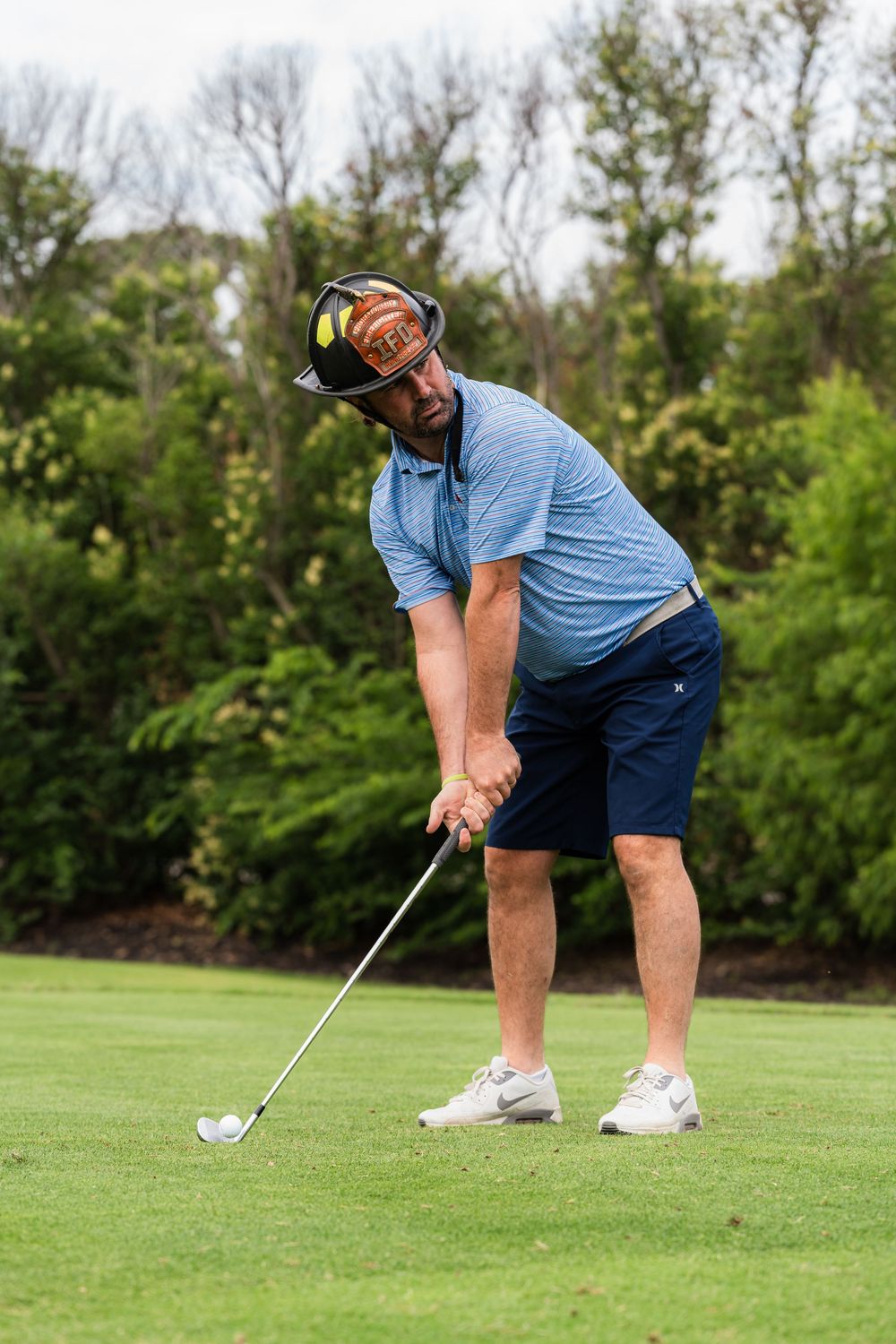 Male golfer wearing a blue shirt and firefighter helmet putting on a green golf course.