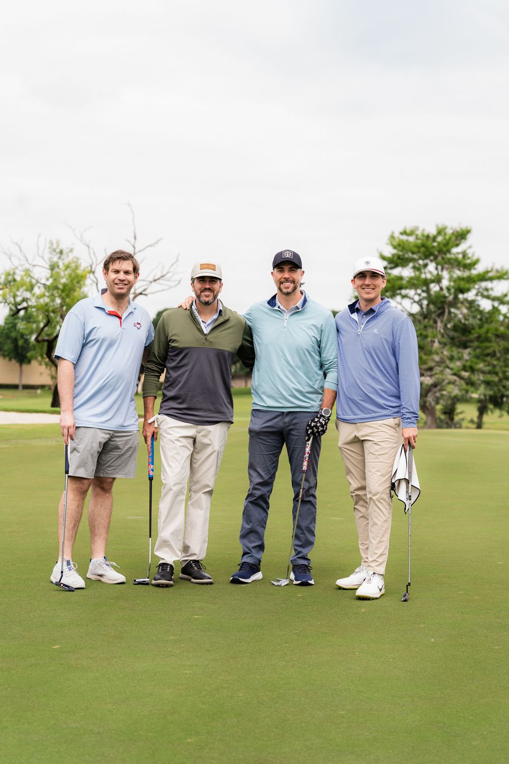 Four male golfers smiling and posing together on a golf course green holding their clubs.