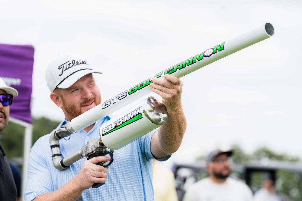 Smiling man in a white Titleist cap aiming a large golf ball cannon at an outdoor event.
