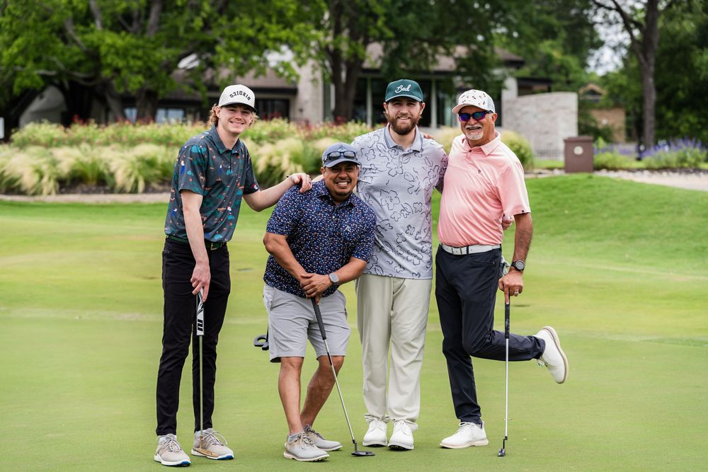 Four smiling golfers posing together on a sunny golf course green during a tournament.
