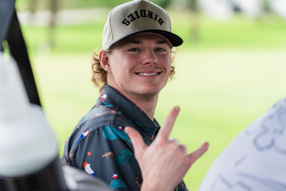 Young man in a trucker cap smiling and making a hand gesture at a golf event.