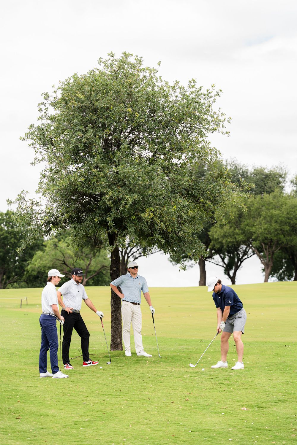 Four golfers on a lush green golf course, one preparing to putt while three others watch nearby.