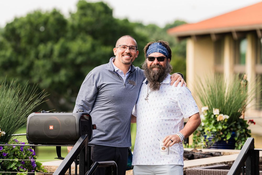 Two smiling men posing together outdoors at a golf event on a sunny day.
