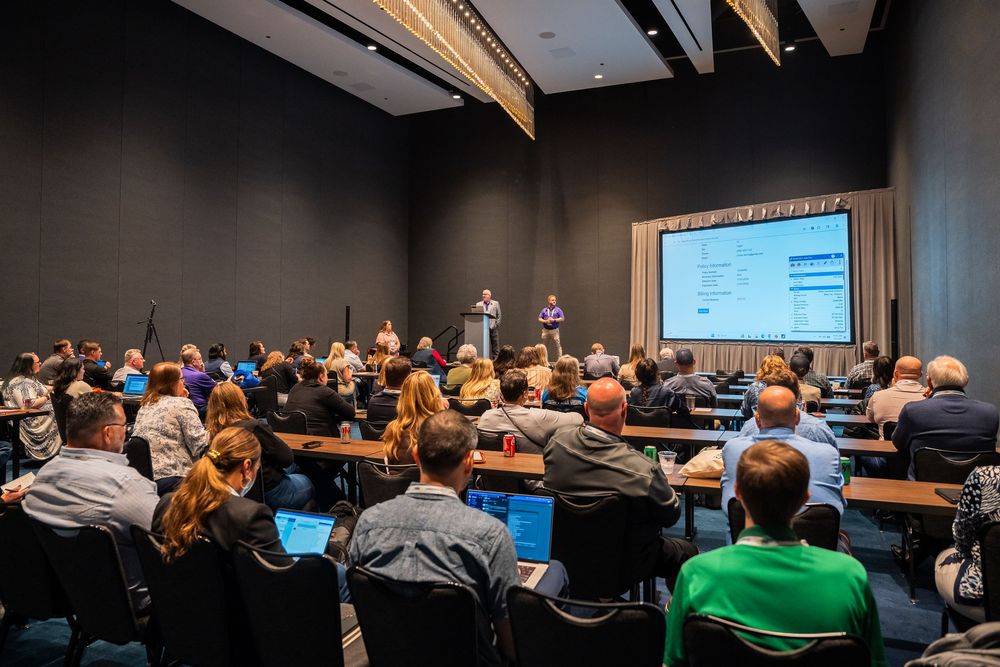 Attendees seated at tables in dark conference room watching a presentation on a large screen.