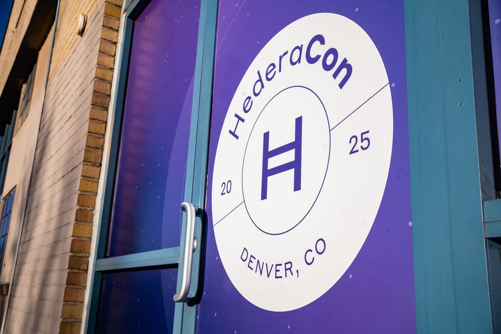 HederaCon 2025 Denver CO event signage displayed on a purple and white circular logo on a glass door.