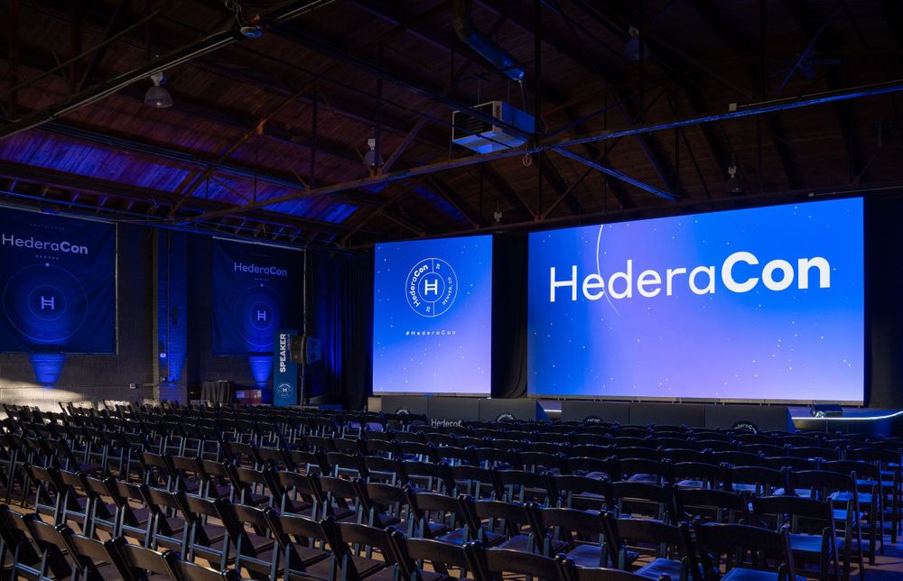 Large HederaCon conference hall with rows of empty chairs facing a massive illuminated stage screen.