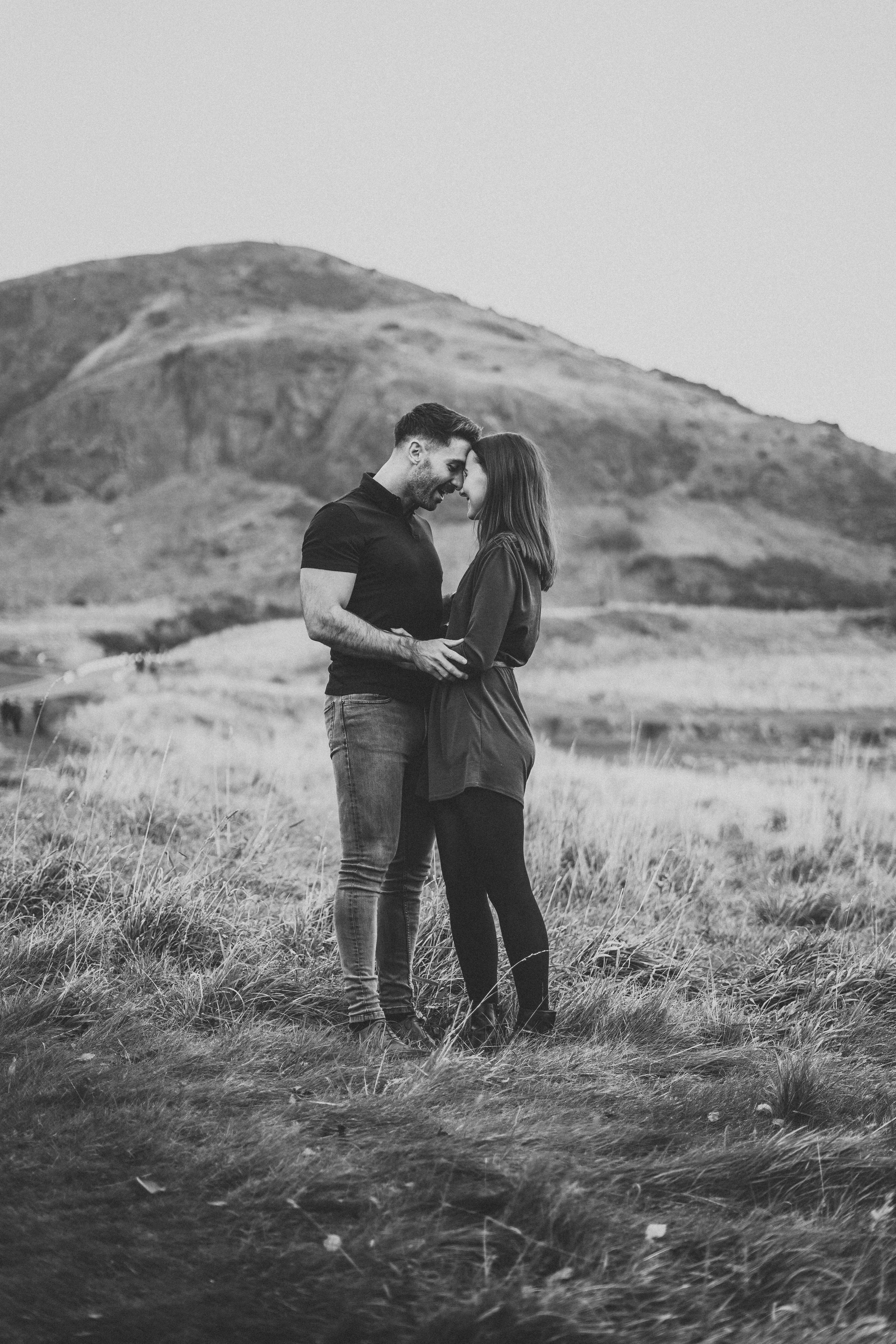 A romantic black and white portrait of a couple standing close together on a hillside.