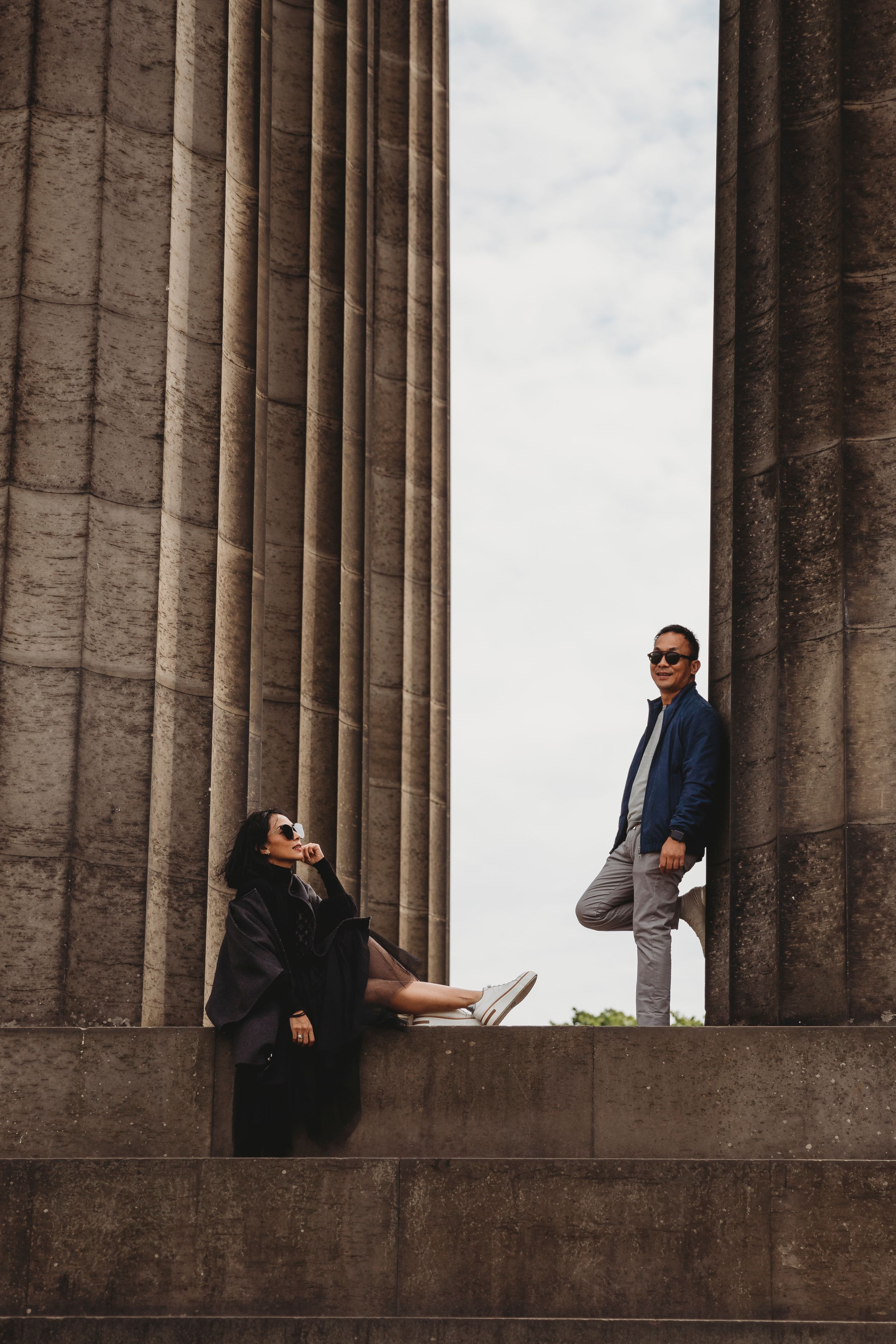 Stylish couple posing between massive stone columns of classical architecture.