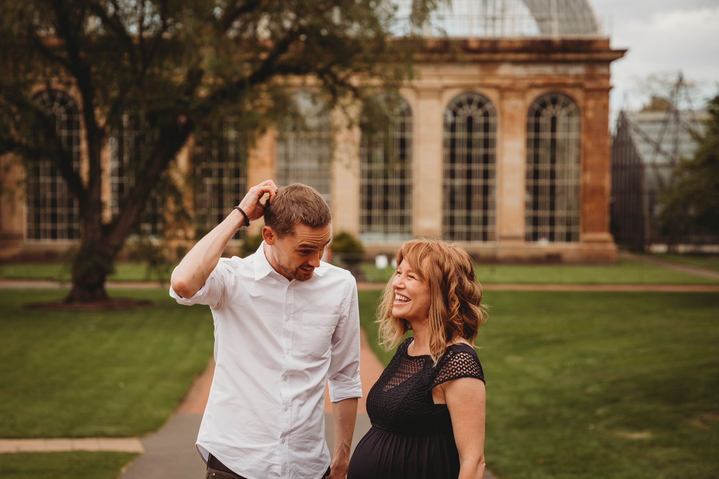 A couple shares a lighthearted moment on a grassy lawn in front of a historic building with arched windows.