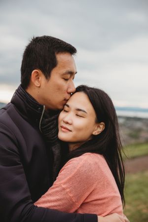 Tender outdoor portrait of couple embracing in coral and dark winter clothing against cloudy sky.