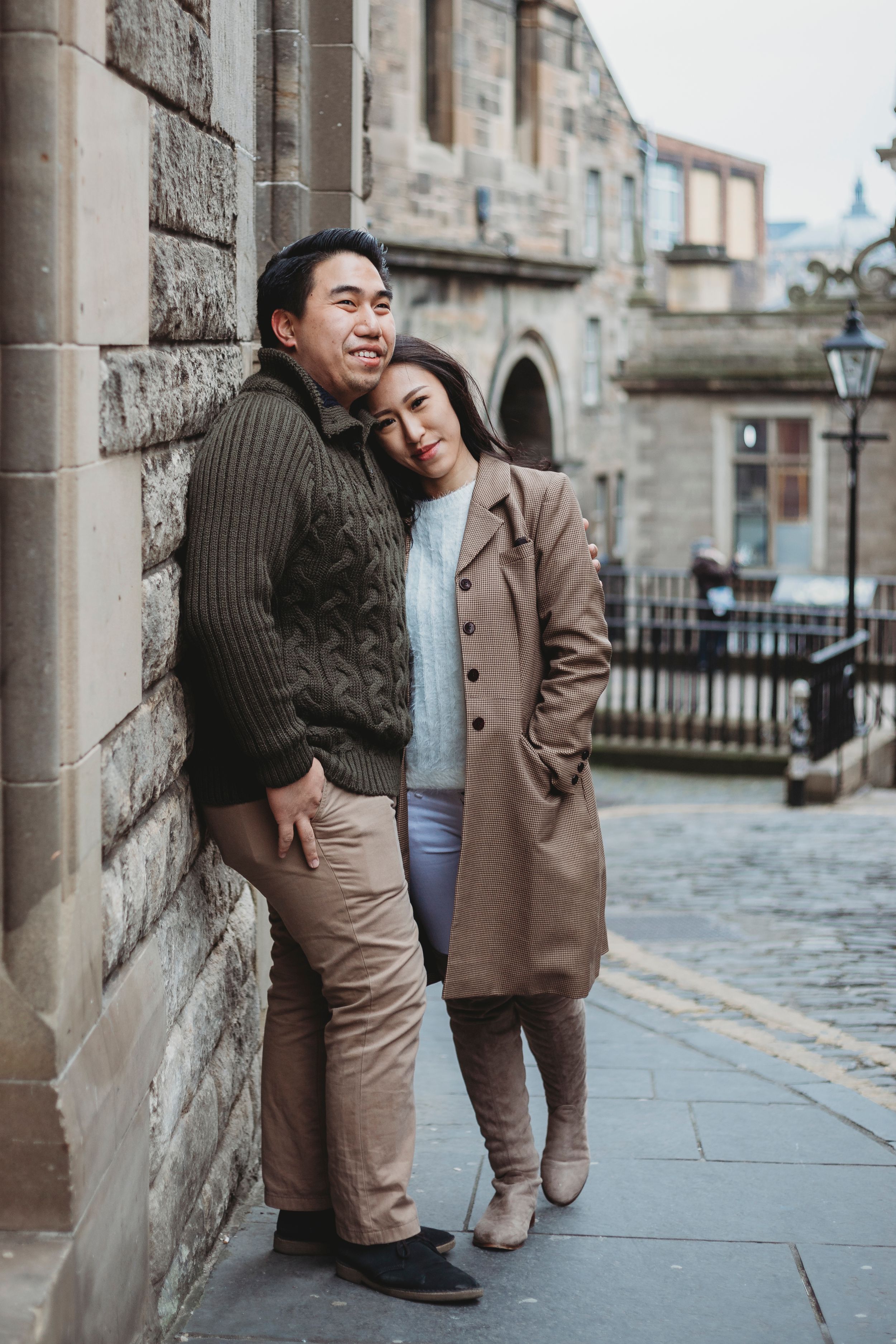 Couple in warm winter clothing embracing against a historic stone building in a European city setting.
