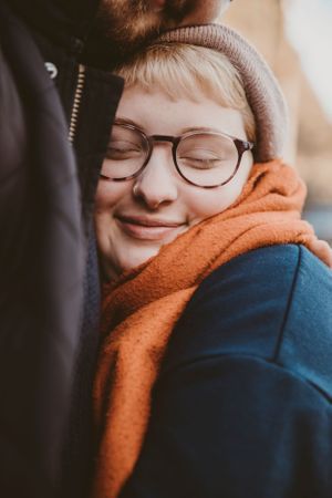 Close-up of someone wearing glasses, beanie, and orange scarf snuggling against another person.