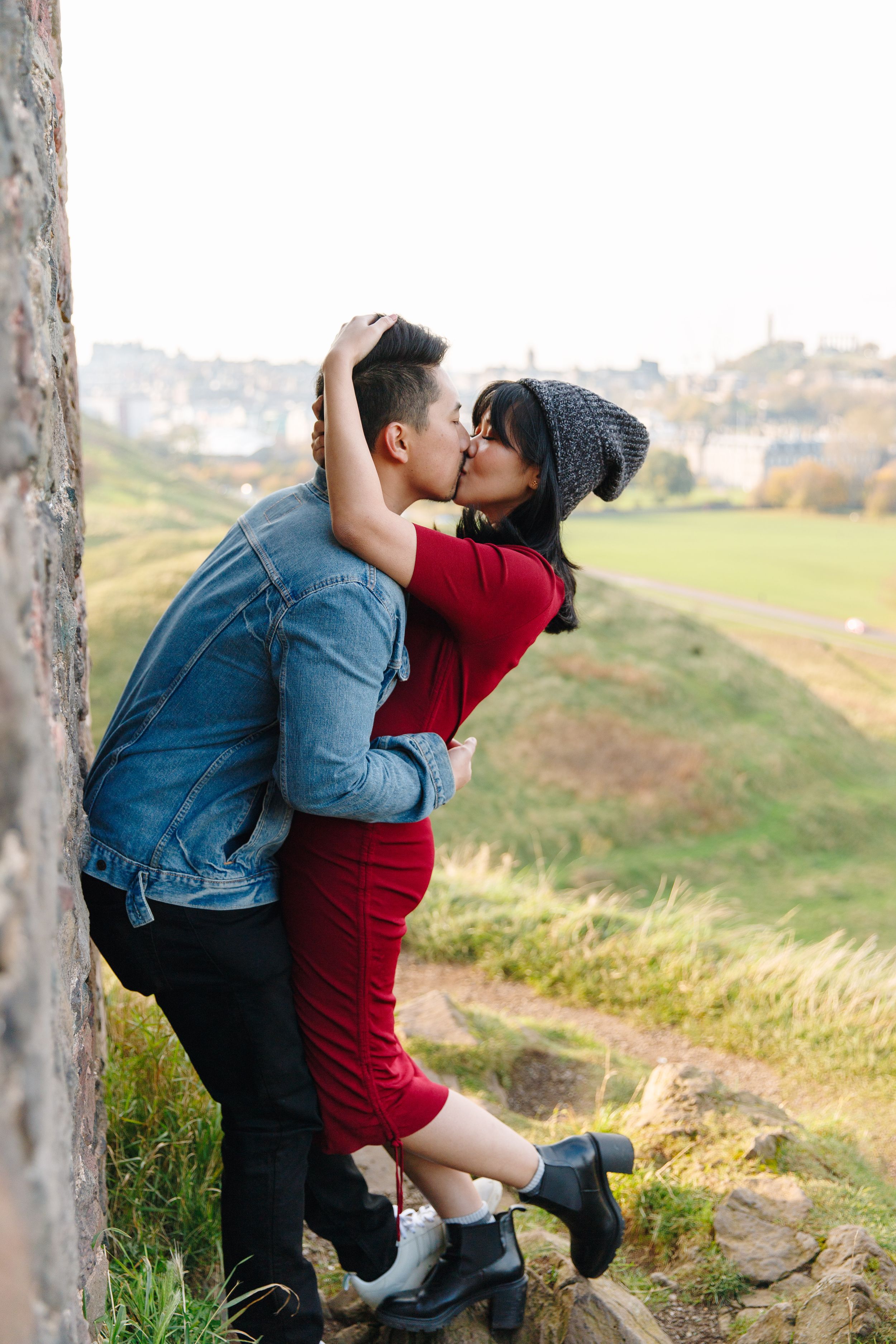 Couple sharing a romantic kiss against stone wall with scenic countryside view in background.