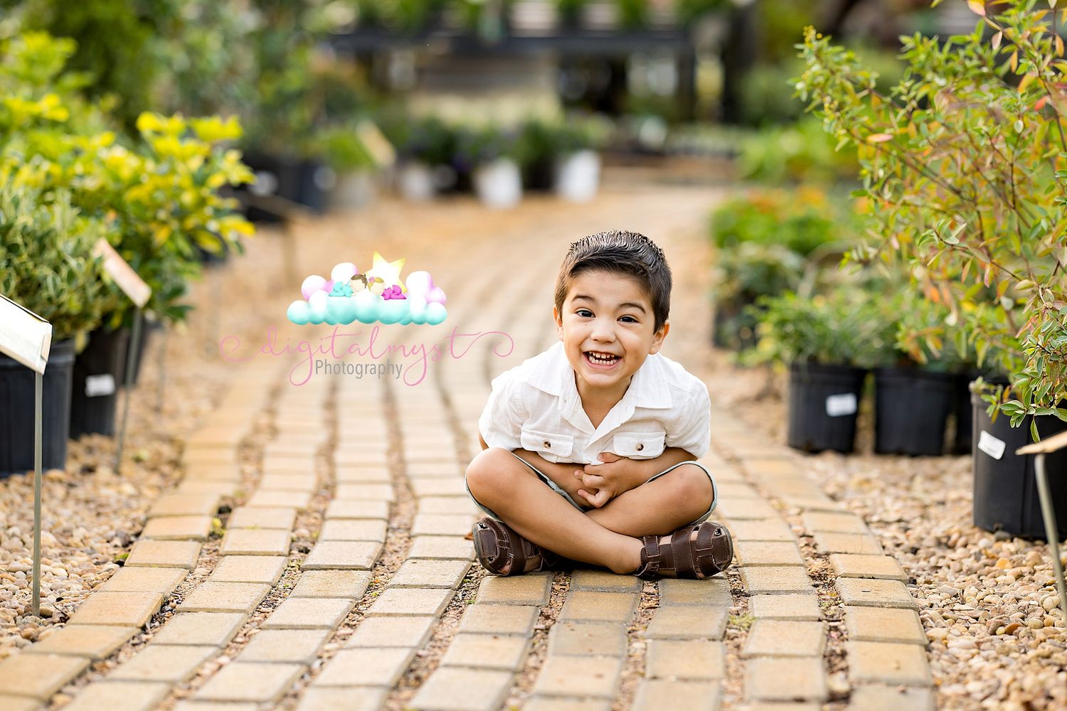 A joyful young boy sits cross-legged on a brick path in a garden nursery, smiling broadly with a birthday cake behind him.