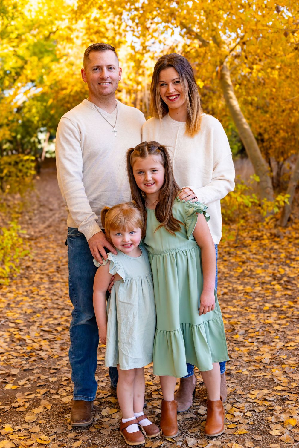 Family of four posing outdoors among golden autumn leaves, parents in cream sweaters with two daughters in sage green dresses.