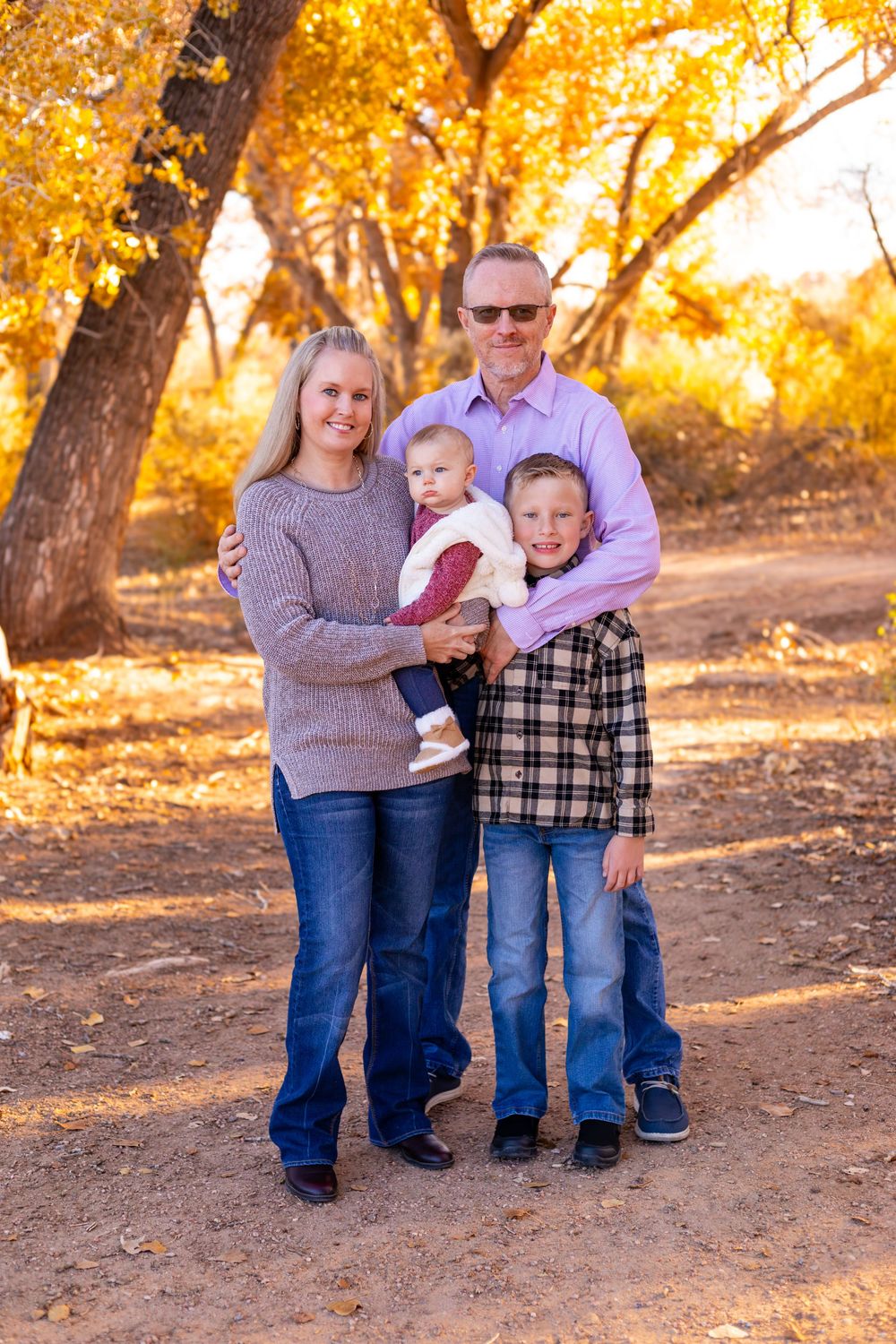 Family of four posing outdoors in autumn park with golden foliage, parents holding baby and young boy smiling at camera.