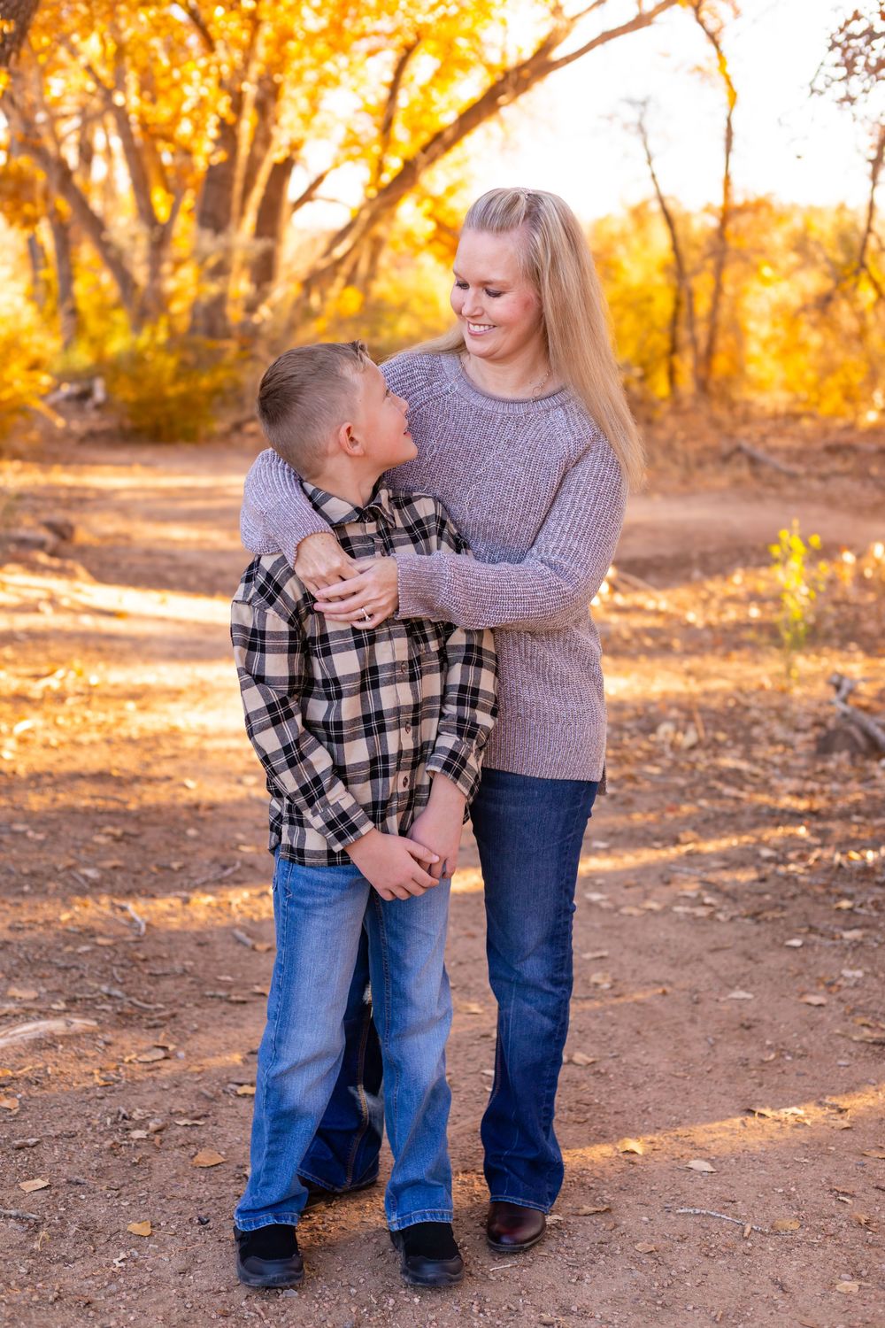 A woman hugs a young boy on a dirt path surrounded by golden autumn trees in a warm fall outdoor portrait.