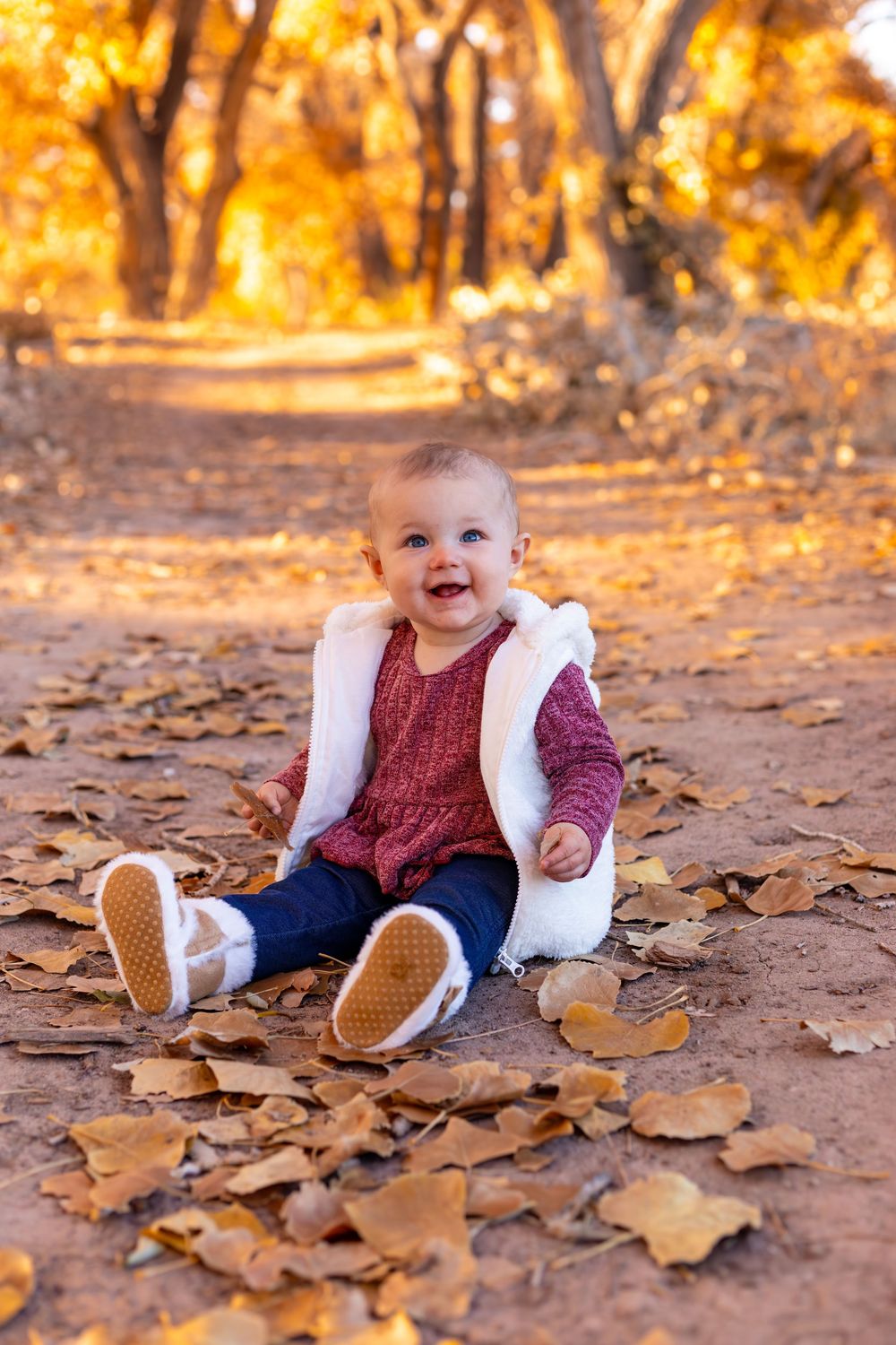 Happy baby girl sitting on fallen autumn leaves in a park, wearing a white vest, red dress, and jeans on a sunny fall day.