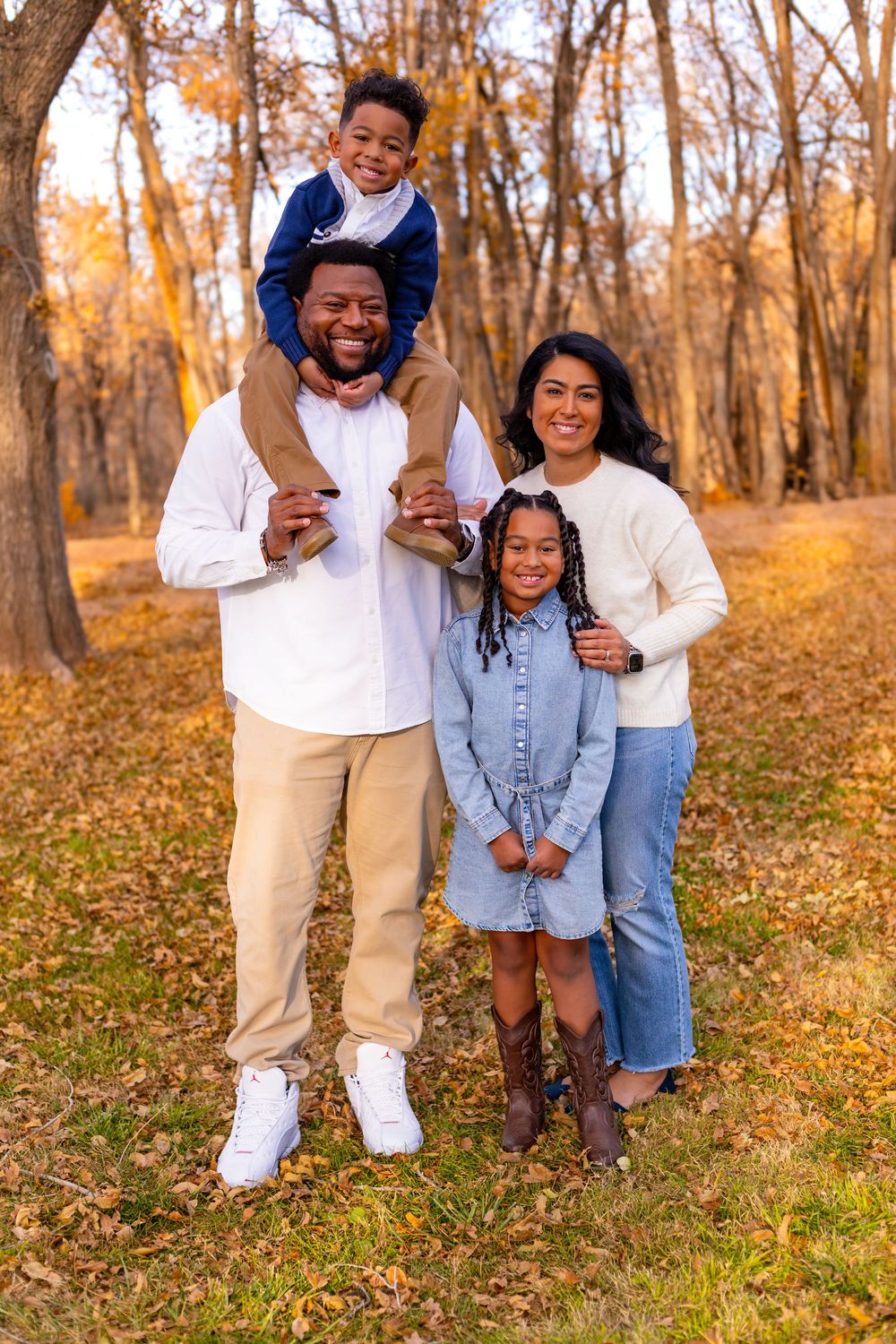 Happy family of four posing in autumn park, father carrying son on shoulders, mother and daughter smiling.