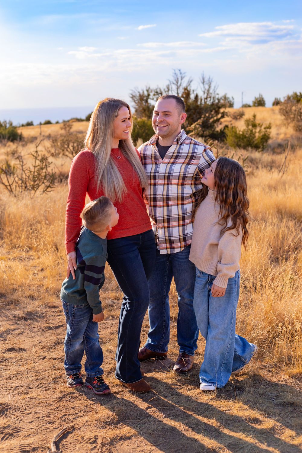 Family of four smiling together outdoors in golden field during sunset, parents with young boy and girl in casual attire.