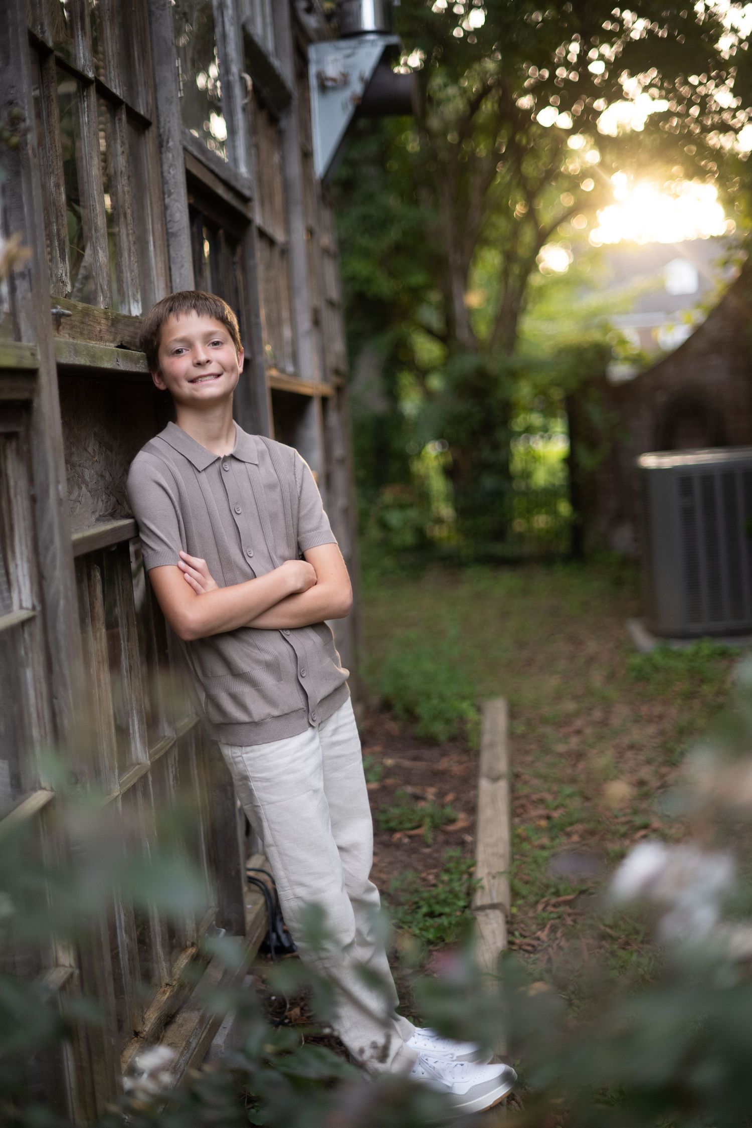 Smiling boy with crossed arms leaning against rustic wooden fence in a lush green backyard at golden hour.