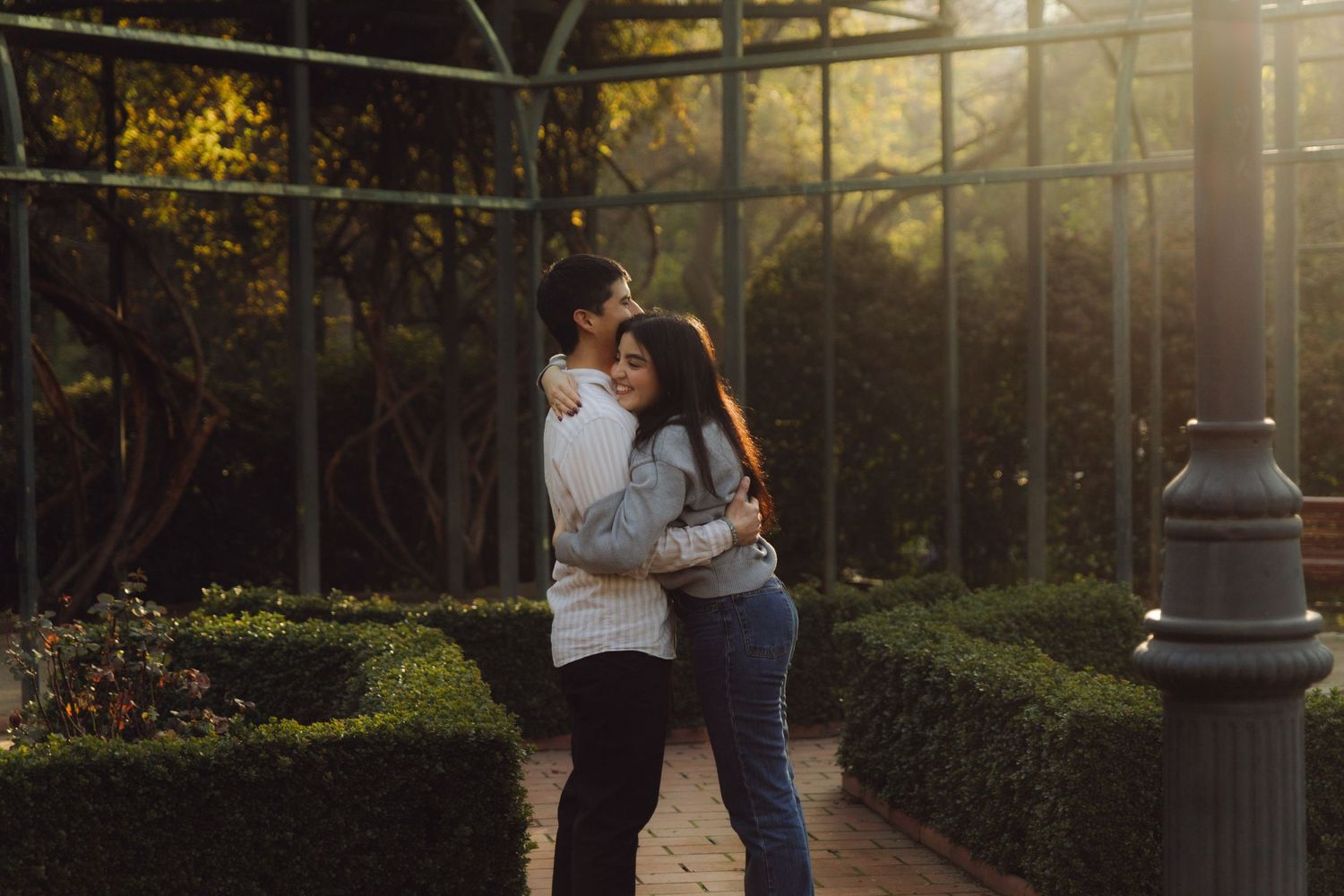 Young couple embracing tenderly in a sunlit garden with lush green hedges and a glass greenhouse backdrop.