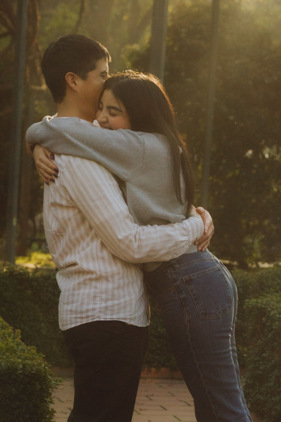Romantic couple sharing a warm hug outdoors in golden sunset light surrounded by green park foliage.