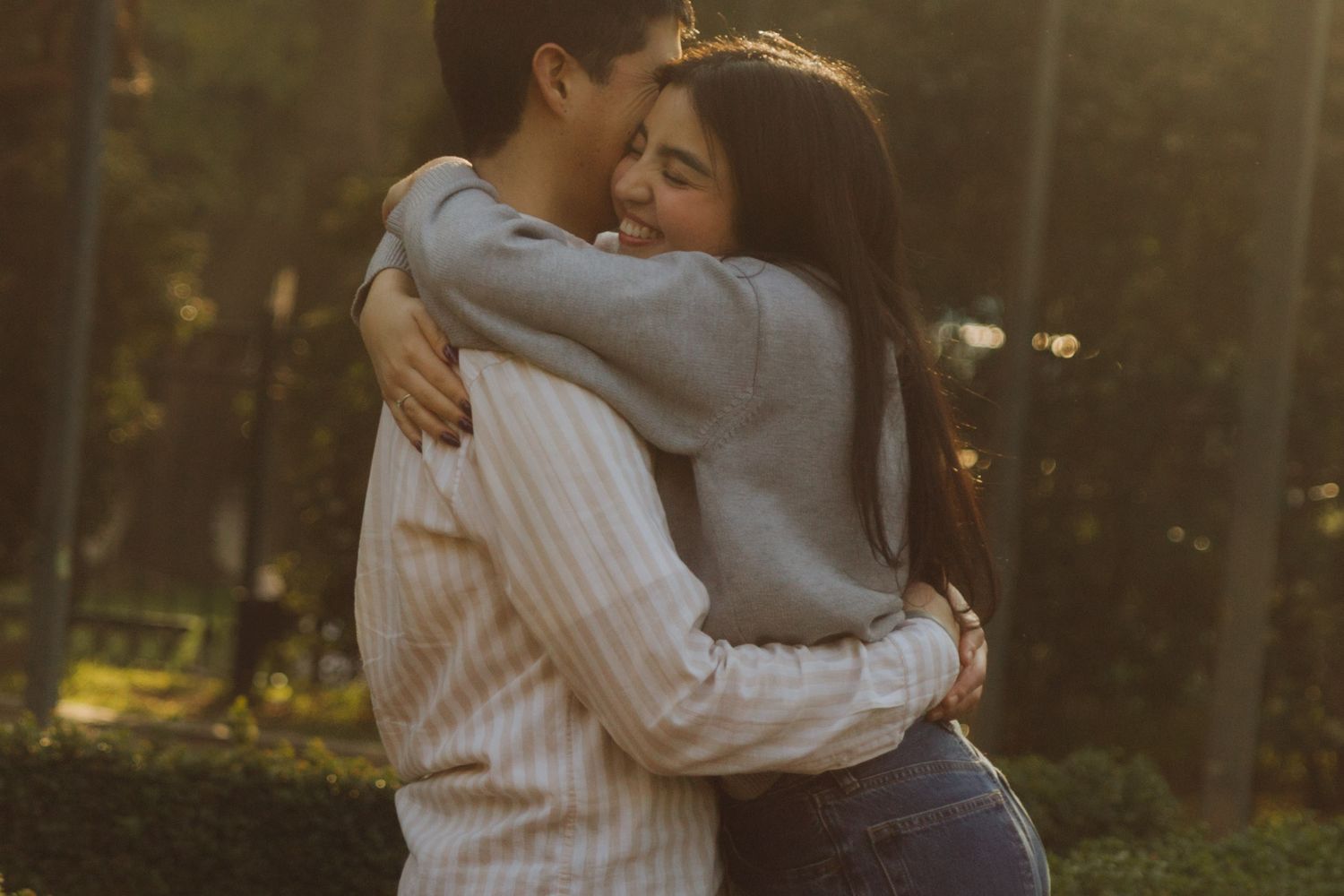 Happy young couple hugging closely in a sun-drenched park, woman smiling joyfully in warm golden light.