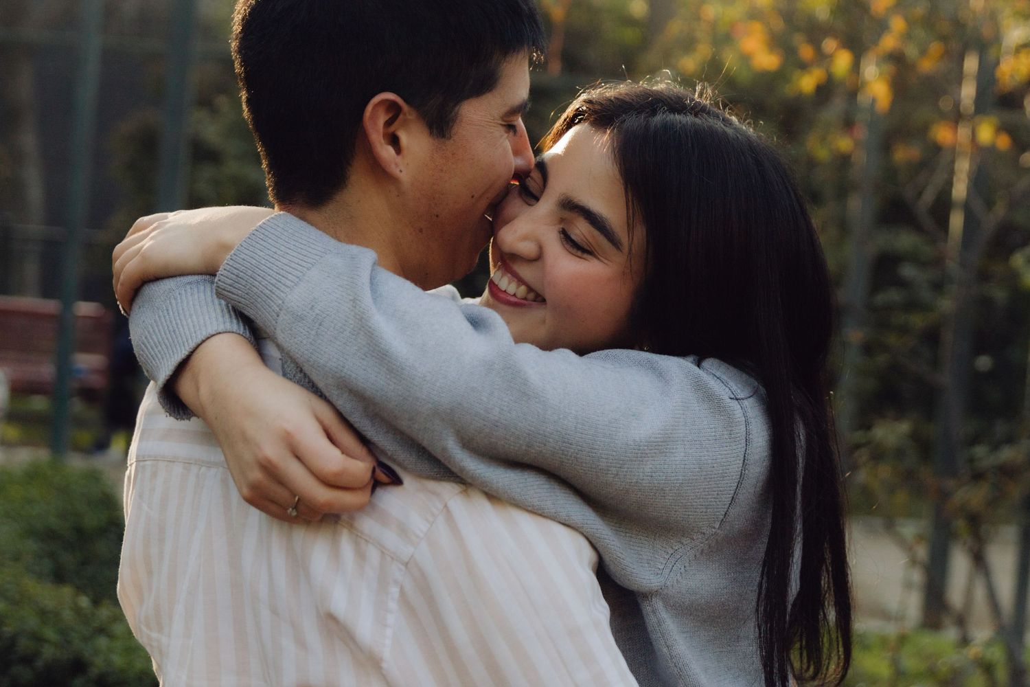 Smiling young woman hugging her partner outdoors in glowing golden hour light with green trees behind.
