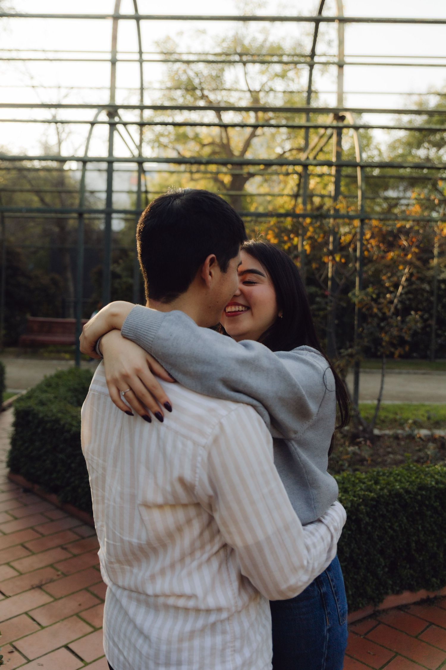 Laughing young woman embracing her partner near a glass greenhouse in warm autumn afternoon sunlight.