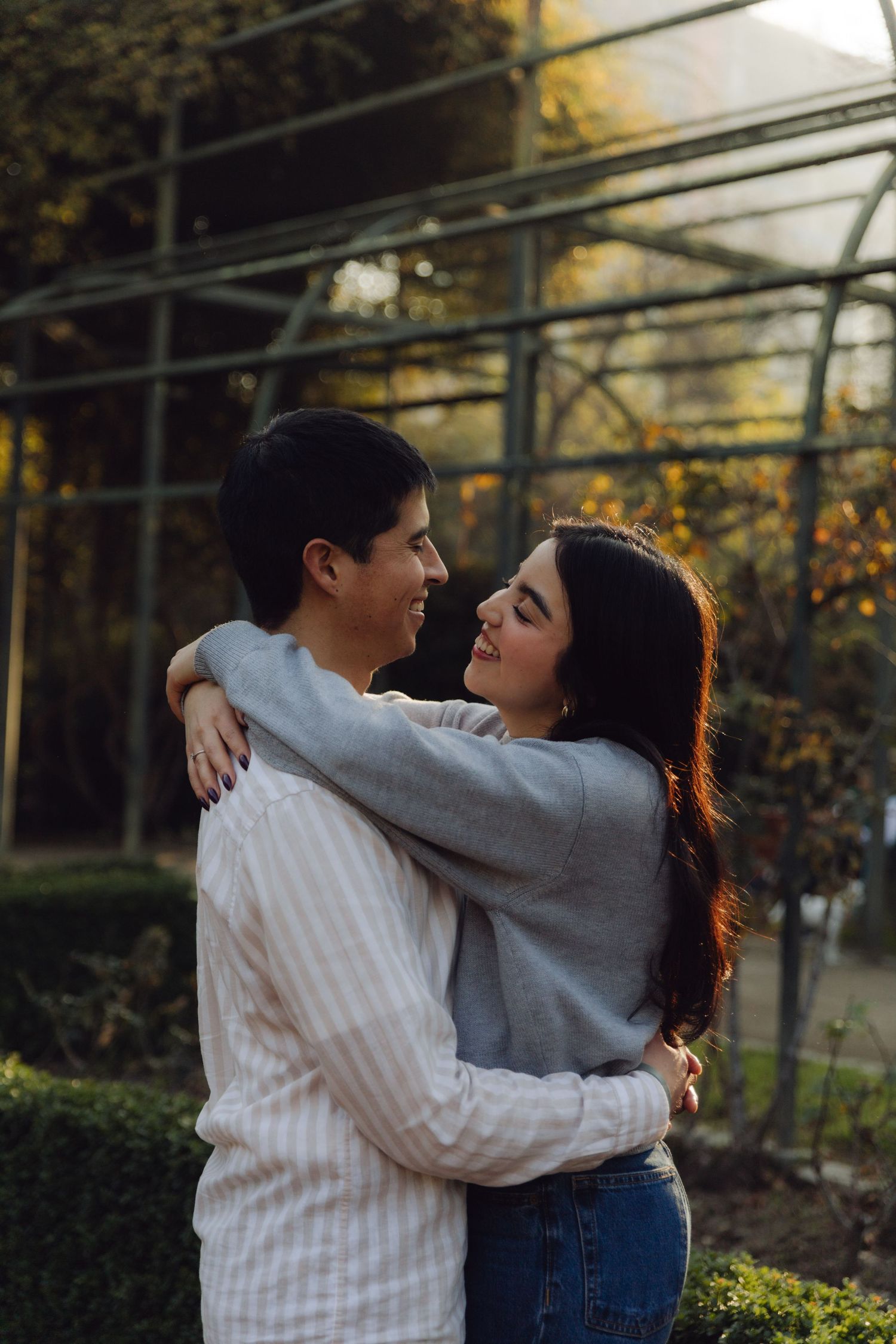 Smiling young couple holding each other close in front of a glass greenhouse in golden evening light.