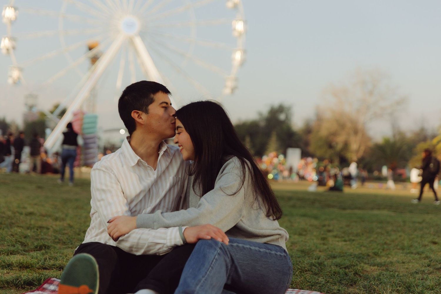 Romantic couple sharing an intimate moment on a picnic blanket with a ferris wheel glowing in the background at dusk.