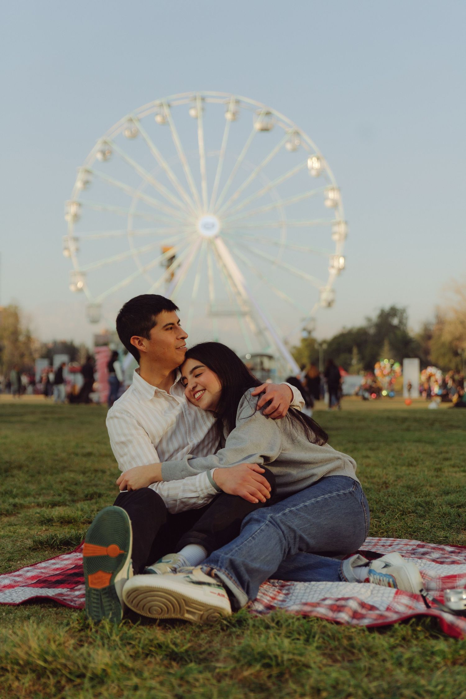 Loving couple sitting together on a red plaid blanket in a park, embracing with a ferris wheel in the background.