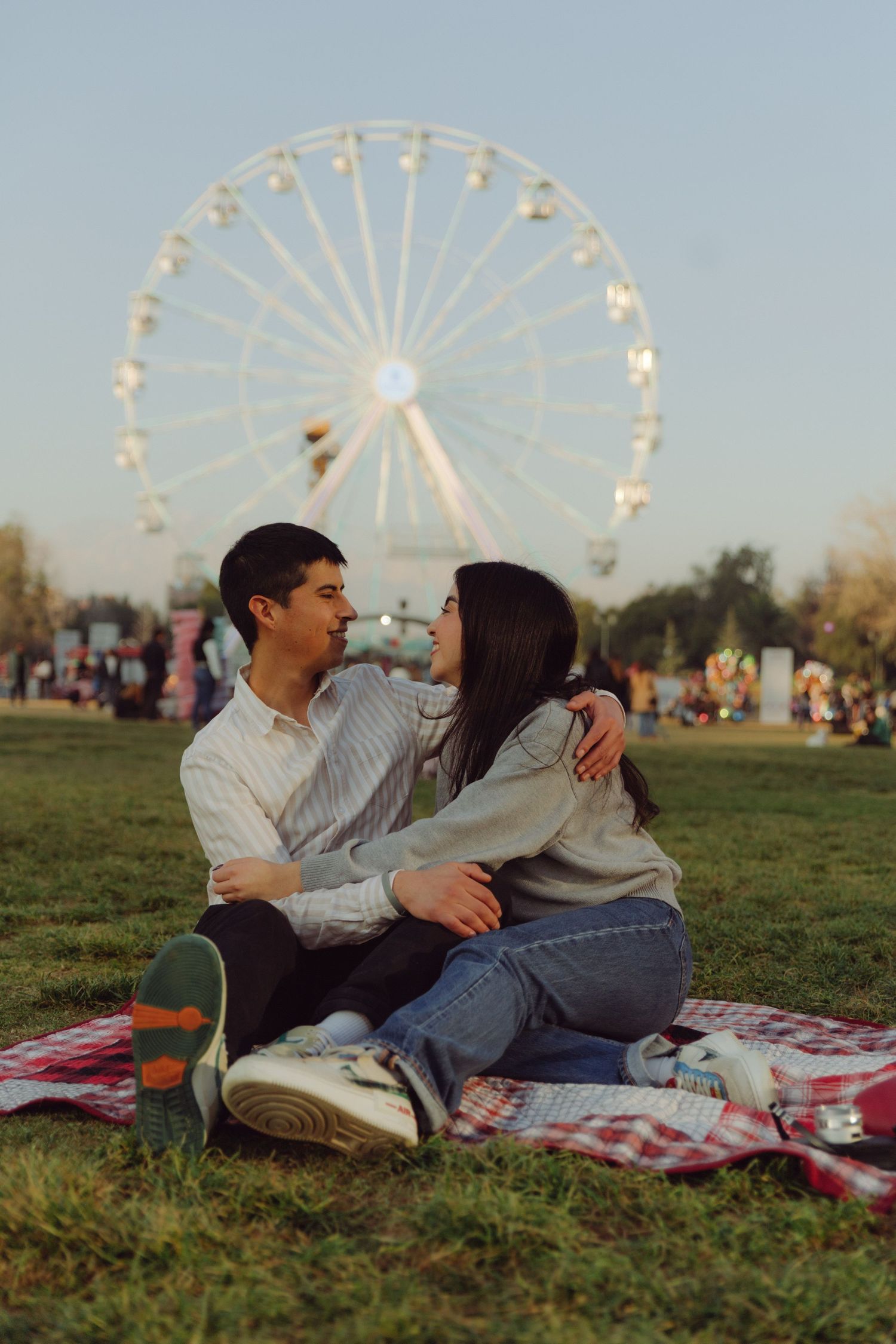 Happy couple sitting on a picnic blanket gazing at each other with a bright ferris wheel visible behind them.