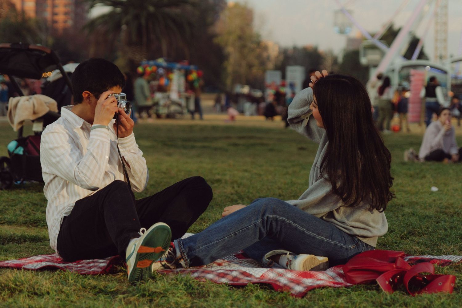 Couple sitting on a picnic blanket in a park, taking photos of each other with film cameras at golden hour.
