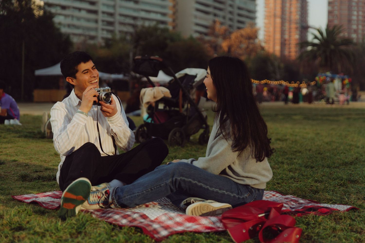 Smiling young man photographing his girlfriend with a film camera while sitting on a blanket in a sunny park.