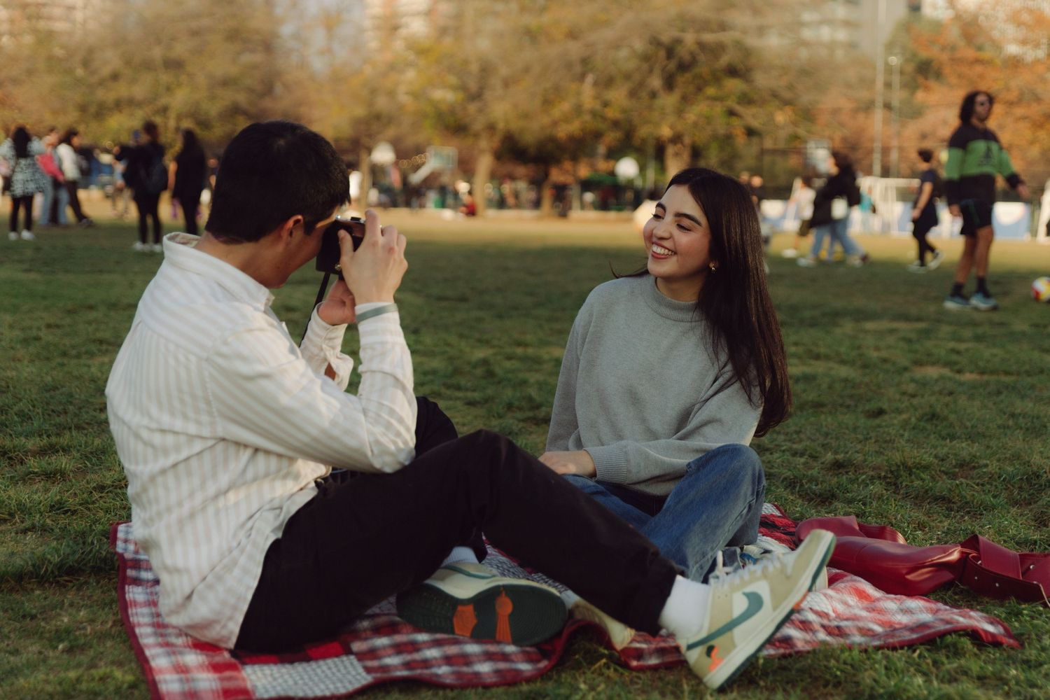 Young woman laughing as her boyfriend photographs her with a film camera on a picnic blanket in a park.