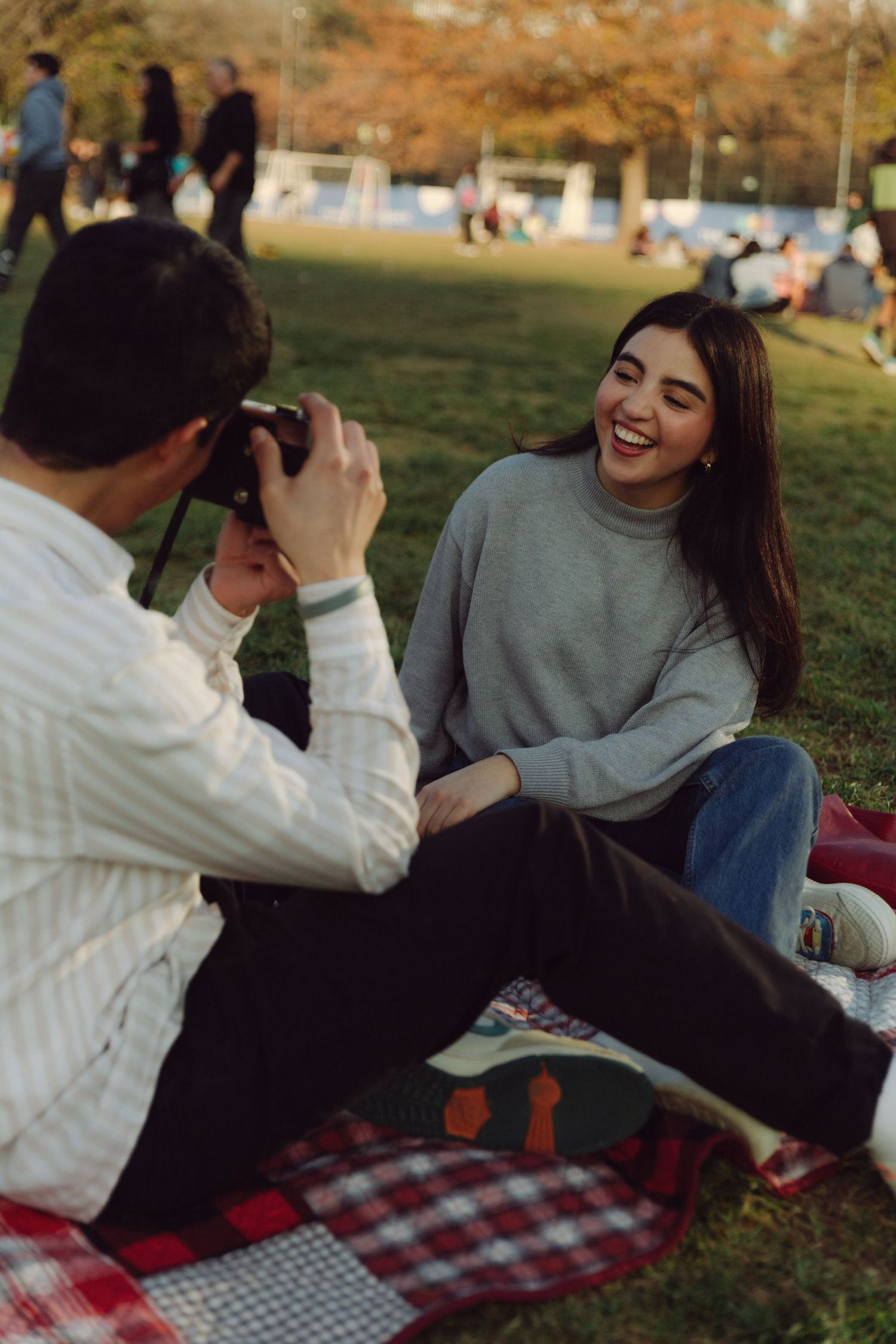 Joyful young woman laughing while her partner photographs her with a vintage camera on a picnic blanket outdoors.