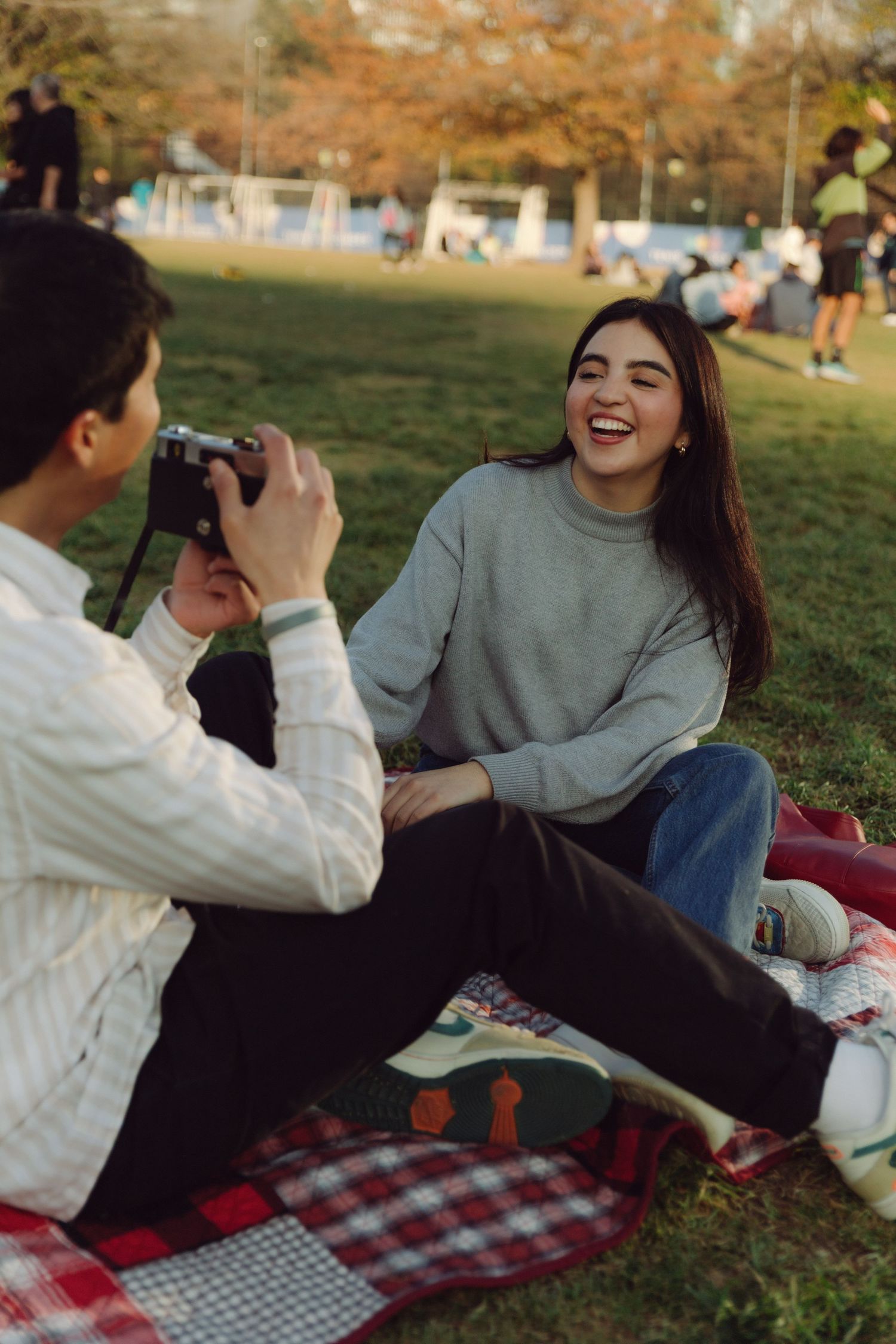 Young woman laughing joyfully as her partner photographs her during a sunny park picnic on a plaid blanket.