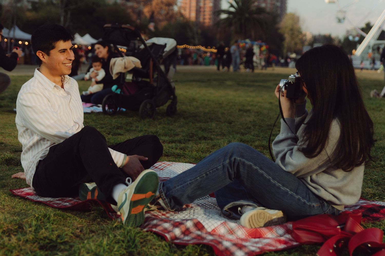 Couple sitting on a picnic blanket in a busy park, woman photographing man with a vintage film camera.