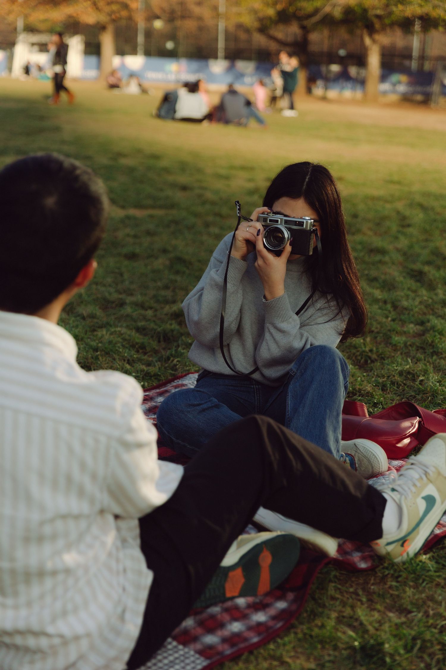Young woman taking photo with vintage camera while sitting on picnic blanket in a sunny park.