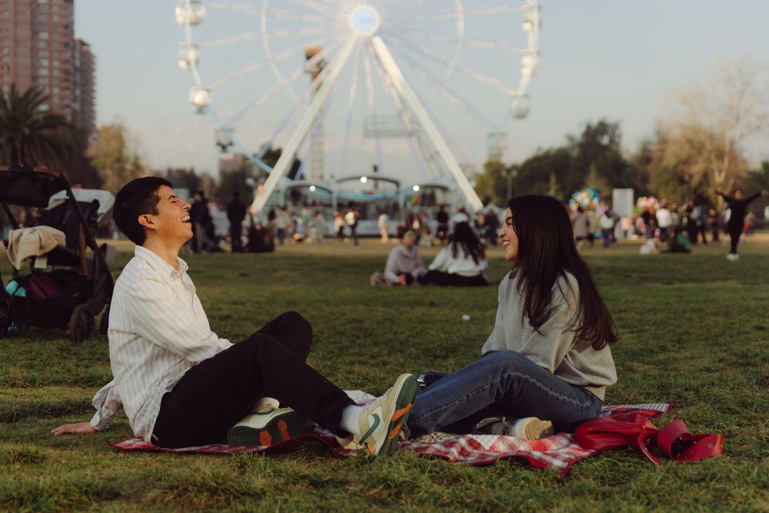 Young couple sitting face-to-face on a picnic blanket with a Ferris wheel and crowd behind them.